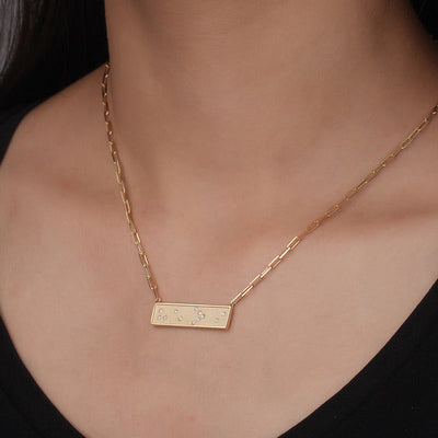 Gold Braille He is Greater Than I necklace with a bar pendant on a person wearing a black top against a neutral background