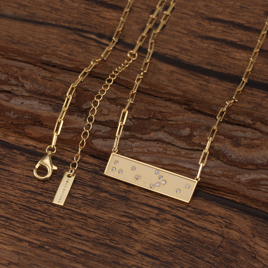 Gold Braille message necklace with a rectangular pendant on a wooden surface