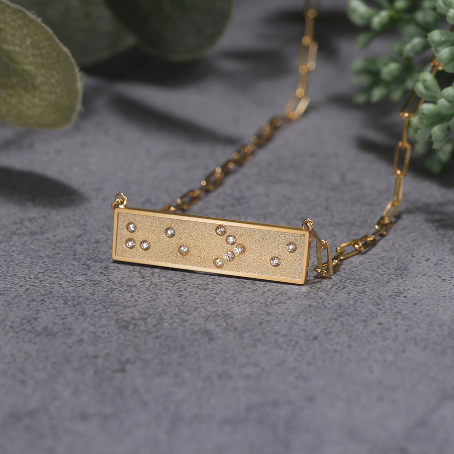 Gold necklace with a rectangular pendant featuring Braille symbols on a gray surface with green leaves in the background.