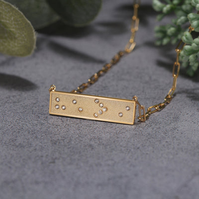 Gold necklace with a rectangular pendant featuring Braille symbols on a gray surface with green leaves in the background.