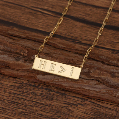 Gold necklace with 'HE>I' pendant on a wooden surface